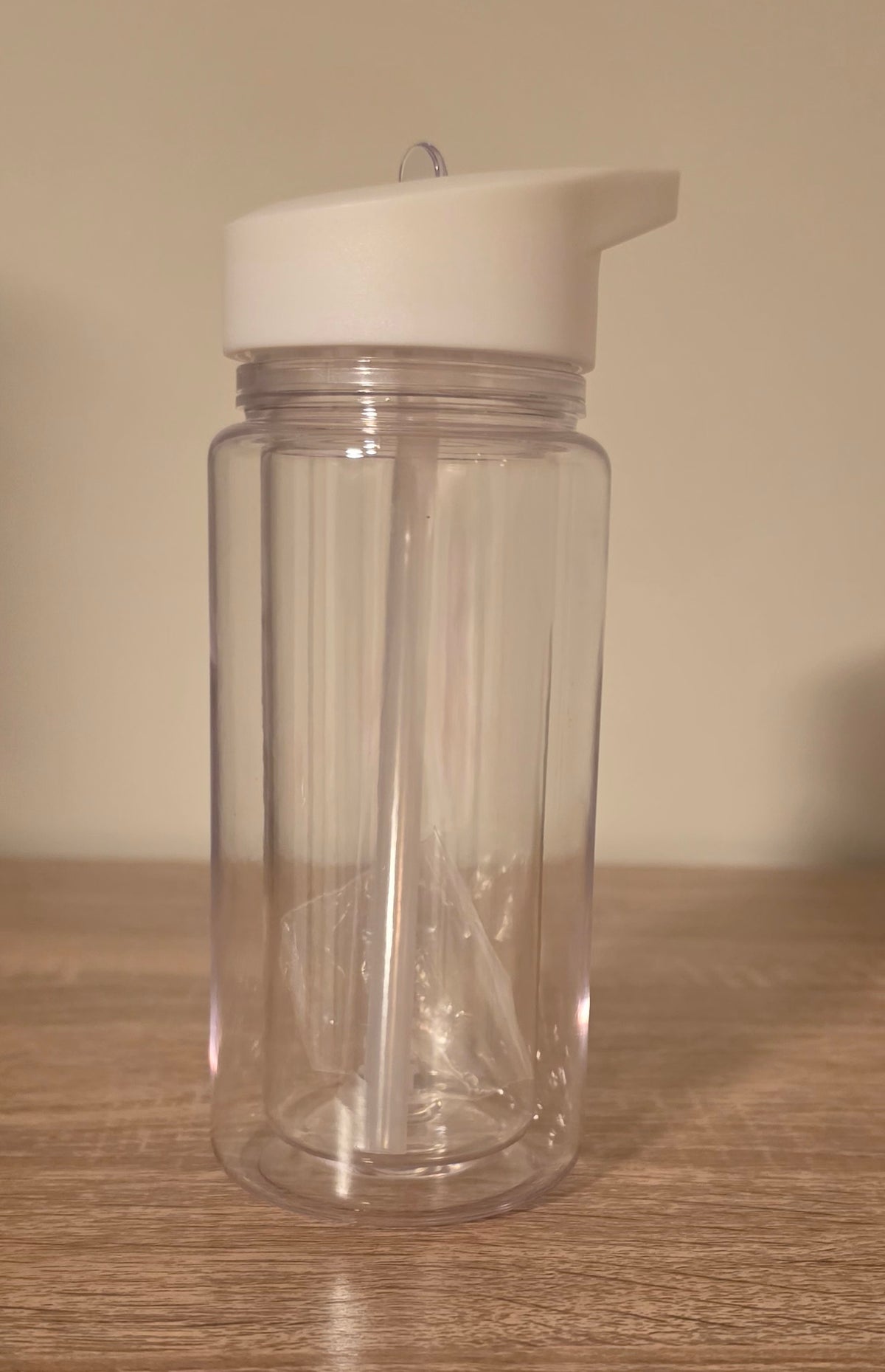 A clear plastic water bottle with a white flip-top lid, shown on a wooden tabletop against a plain beige wall.
BH Creations By Mel