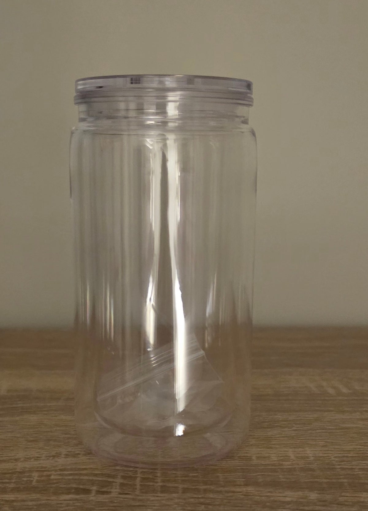 A clear plastic jar with a ribbed wall and a matching clear screw-on lid, standing on a wooden surface against a plain beige wall.
BH Creations By Mel
