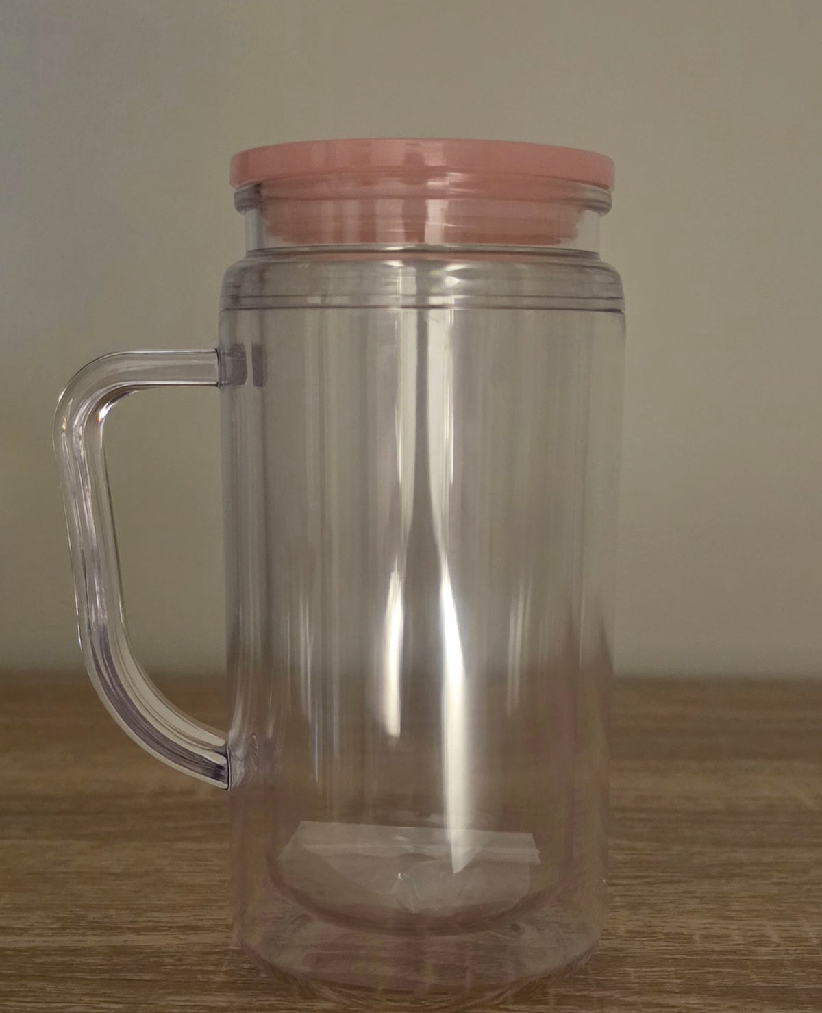 A clear plastic handled mug with a light pink lid, shown on a wooden surface against a plain beige wall.
BH Creations By Mel