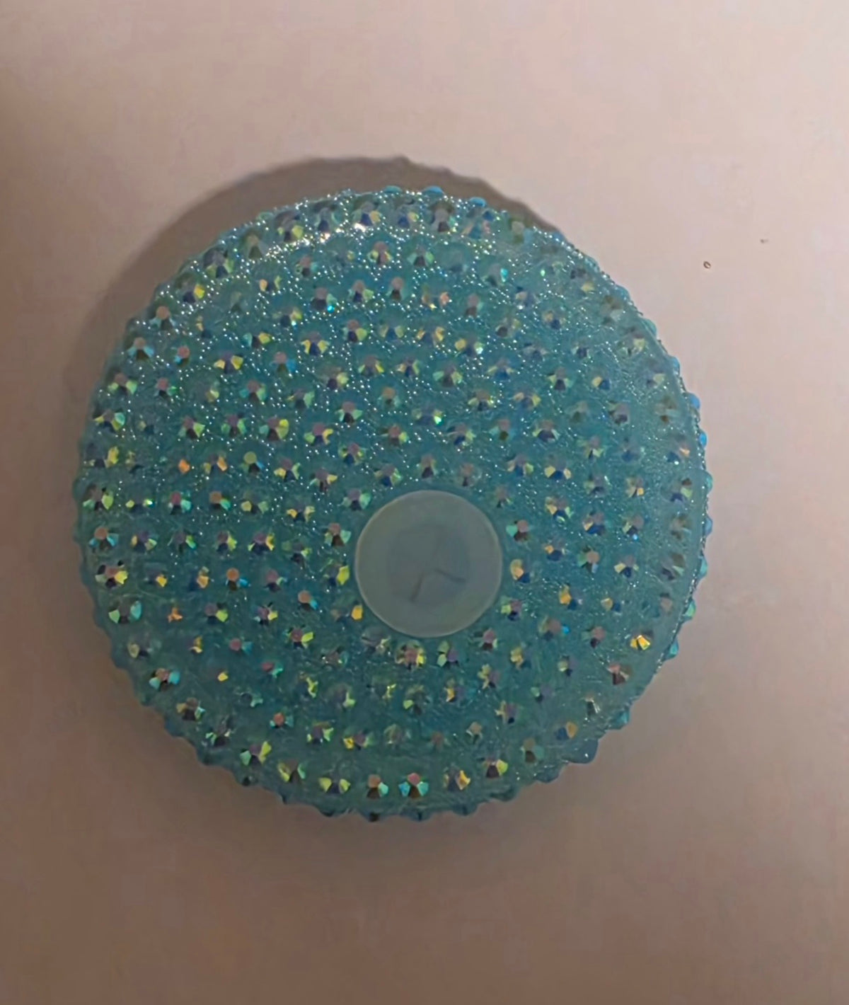 A round, textured light blue plastic item covered in small iridescent rhinestones, with a translucent circular center, shown on a plain beige background.
BH Creations by mel