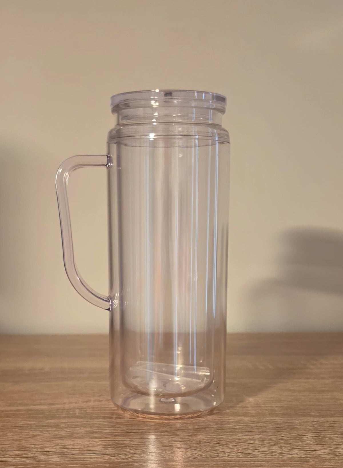 A clear plastic double-walled tumbler with a handle, shown empty on a wooden tabletop against a plain beige wall.
BH Creations By Mel