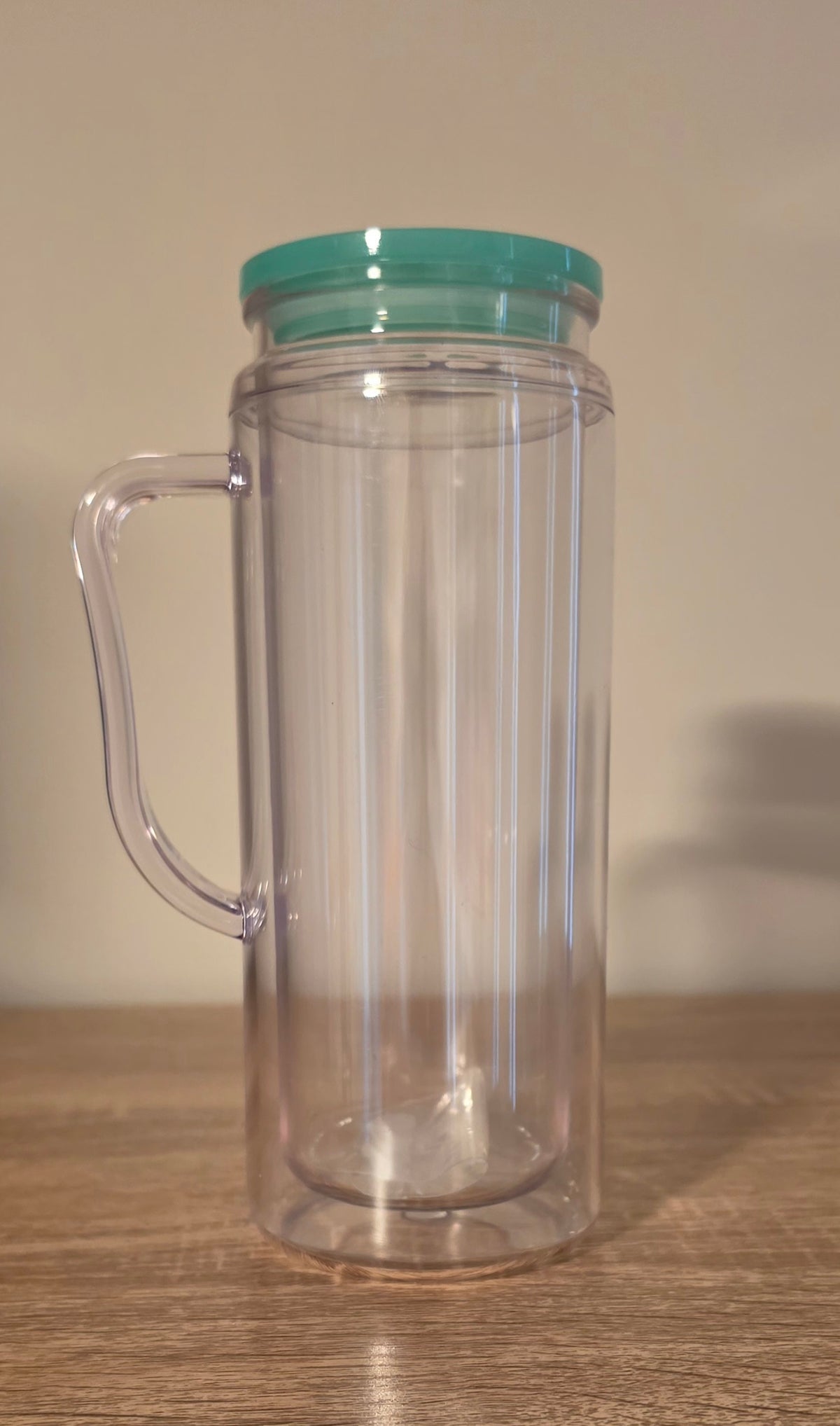 A clear plastic double-walled tumbler with a handle and a mint green lid, standing on a wooden surface against a plain beige wall.