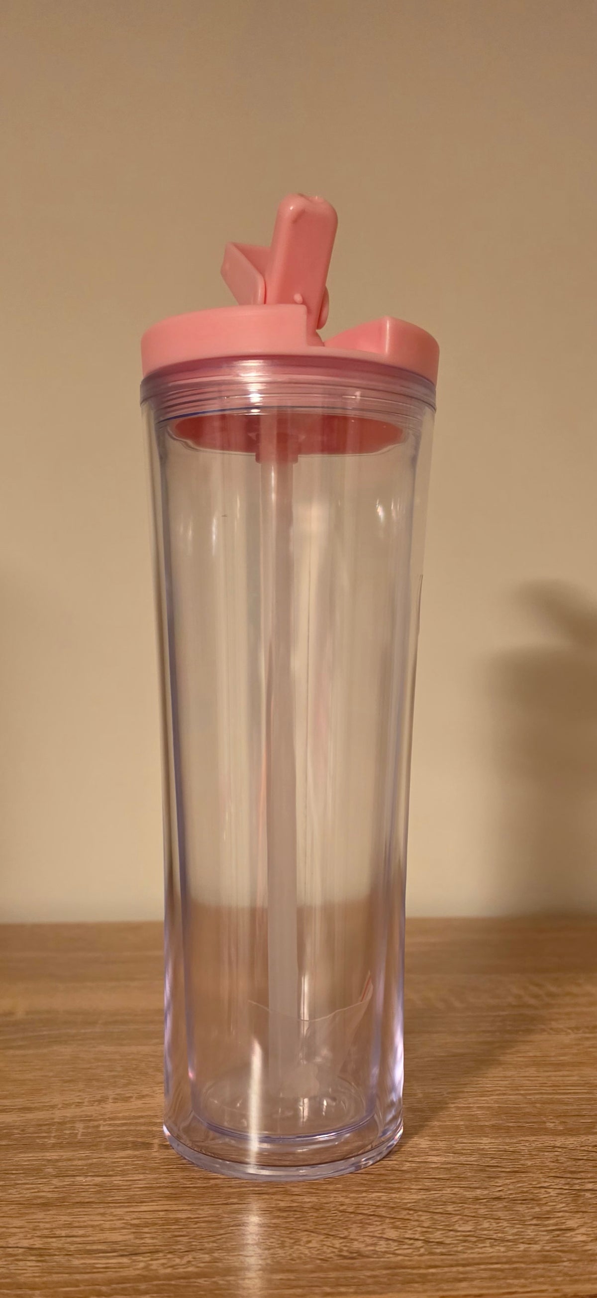 A tall, clear plastic travel tumbler with a pink flip-top lid and straw, standing on a wooden surface against a plain beige wall.
BH Creations by mel