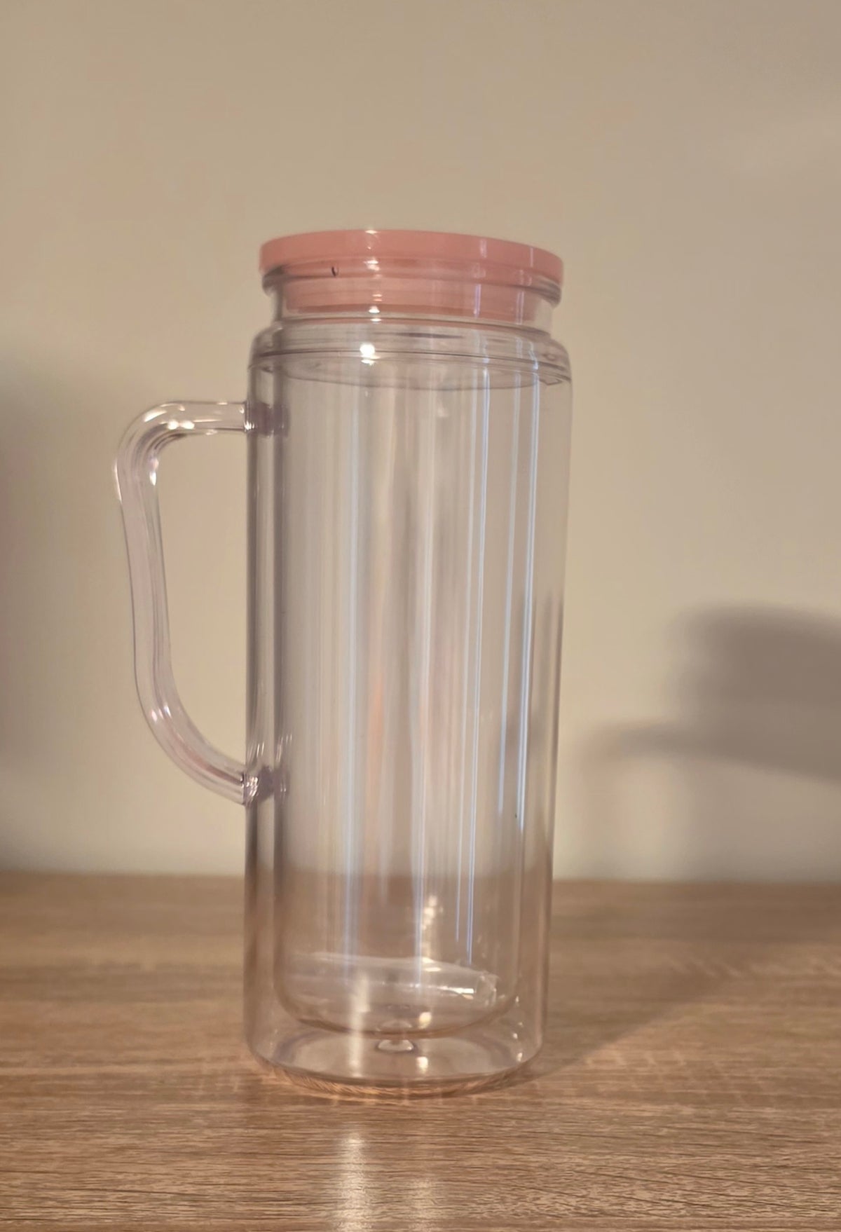 A clear double-walled glass tumbler with a pink lid and integrated handle, sitting on a wooden surface against a plain beige wall.
BH Creations By Mel