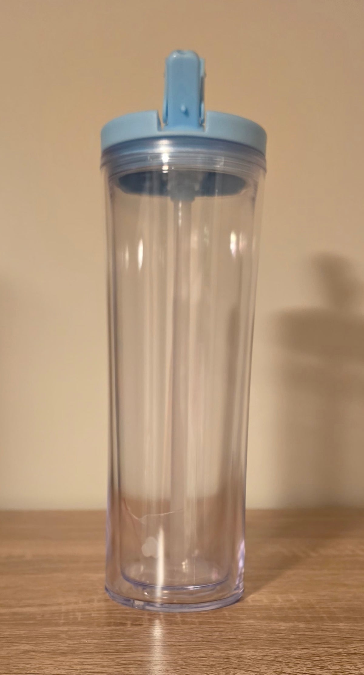 A clear plastic travel tumbler with a light blue flip-top lid, shown on a wooden surface against a beige wall.
BH Creations by mel