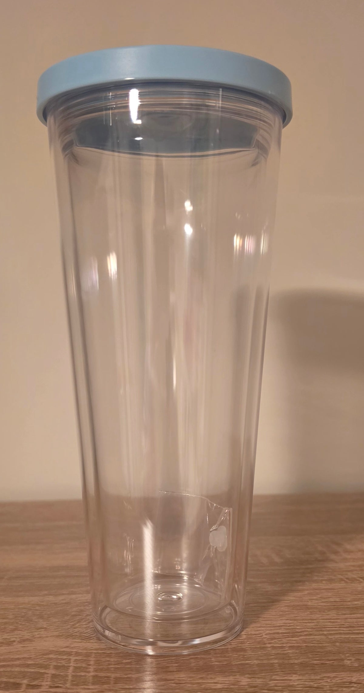 A clear plastic travel tumbler with a light blue lid, standing on a wooden surface against a plain beige wall.