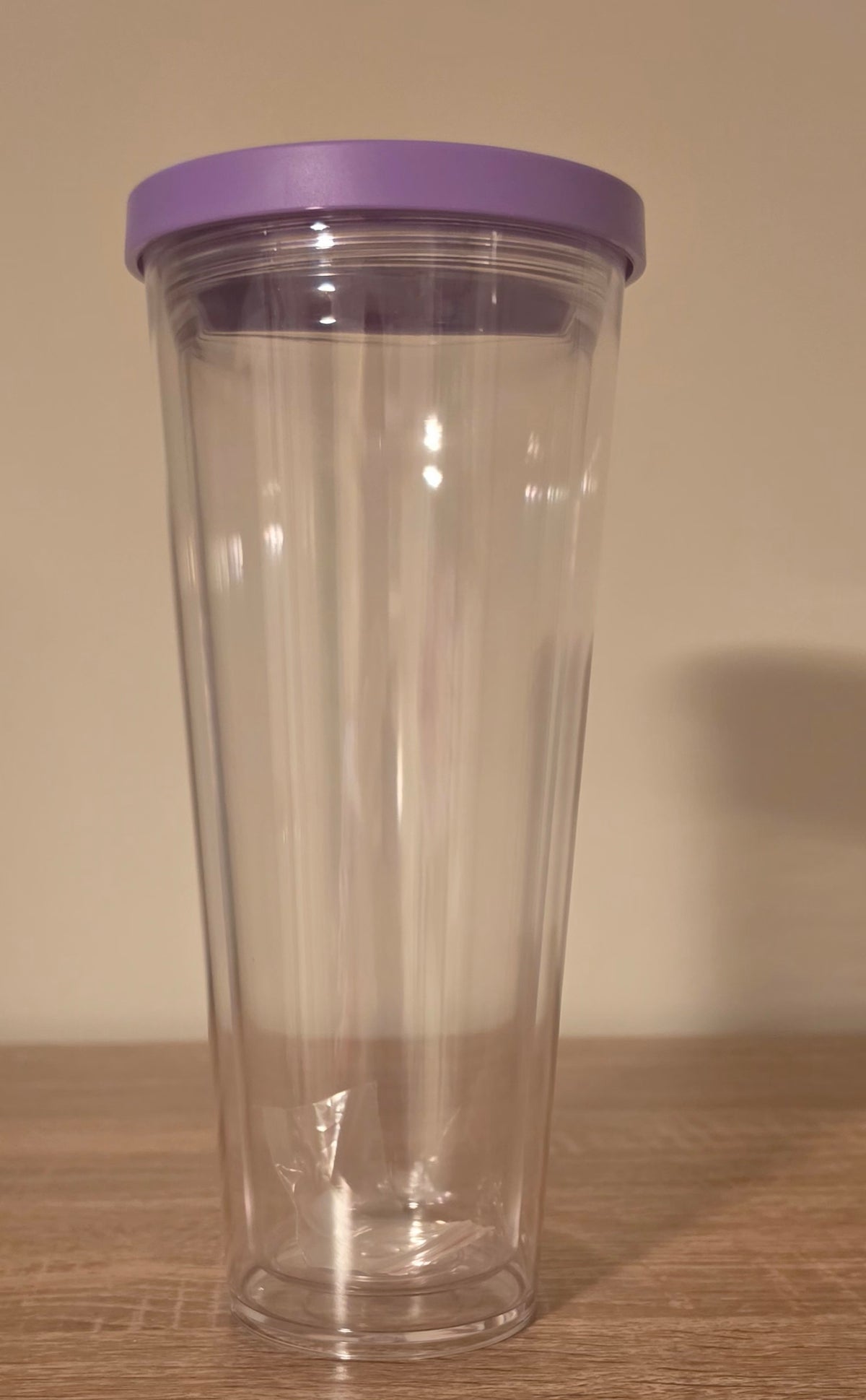 A Clear plastic travel tumbler with a purple screw on lid, standing on a wooden surface against a plain beige wall.
BH Creations By Mel