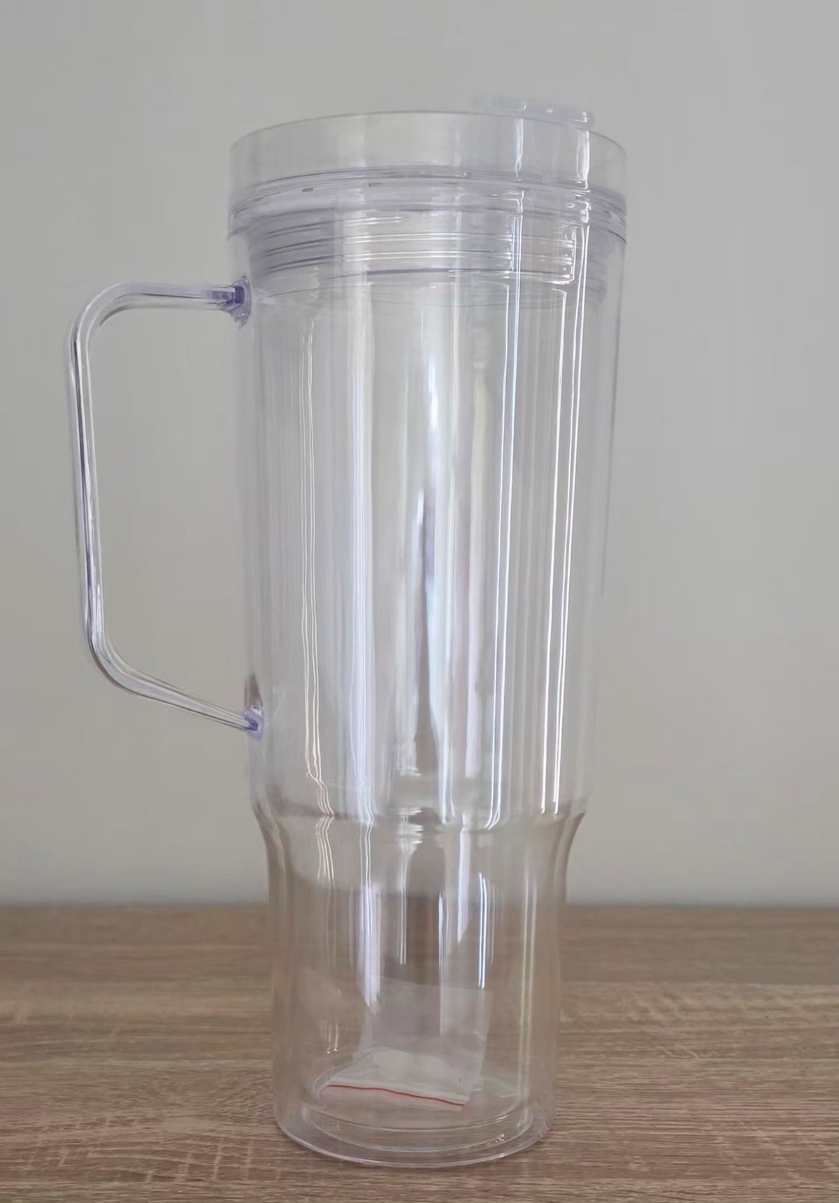 A clear plastic travel tumbler with a handle and lid, shown empty on a wooden surface against a plain light gray wall.
BH Creations By Mel